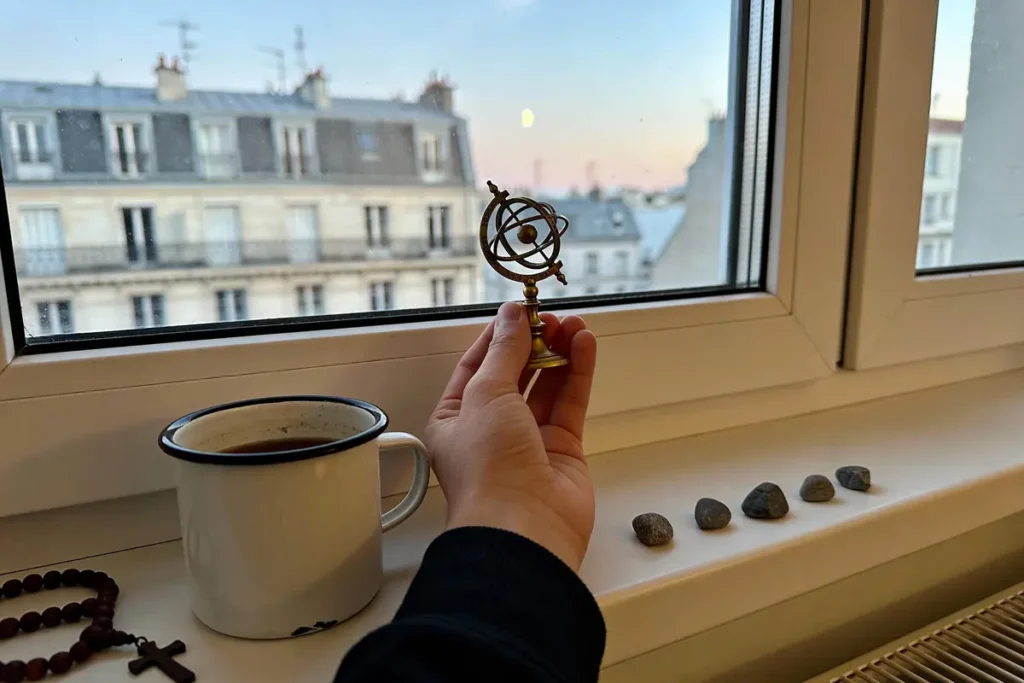 Main tenant astrolabe miniature, tasse de café, perles de chapelet, pierres, vue sur immeubles parisiens.