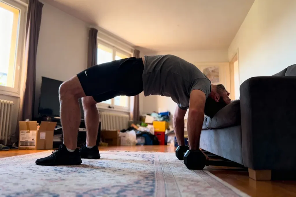 Homme faisant des exercices de pont avec haltères, pieds au sol, tête sur canapé, intérieur salon lumineux.