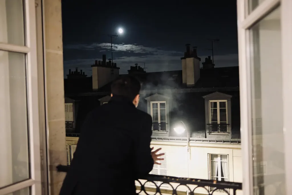 Homme regardant la nuit depuis un balcon, pleine lune visible, toits et cheminées urbaines côtières.