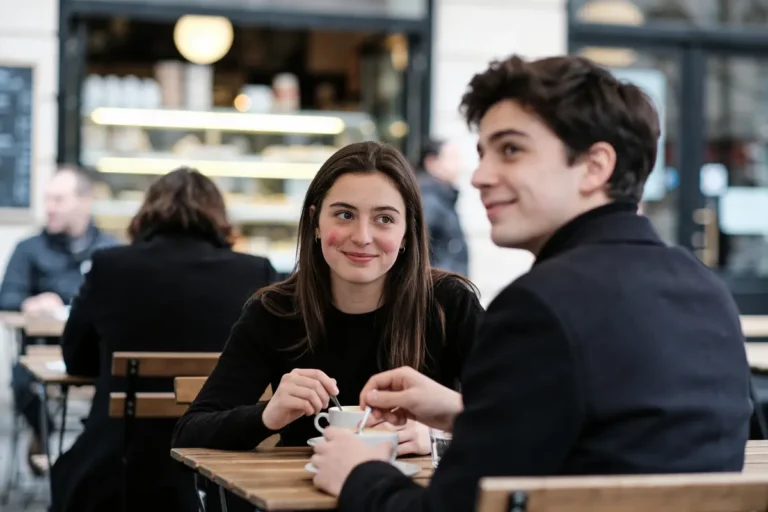 Deux personnes souriantes prennent un café ensemble dans un café animé avec des tables en bois.
