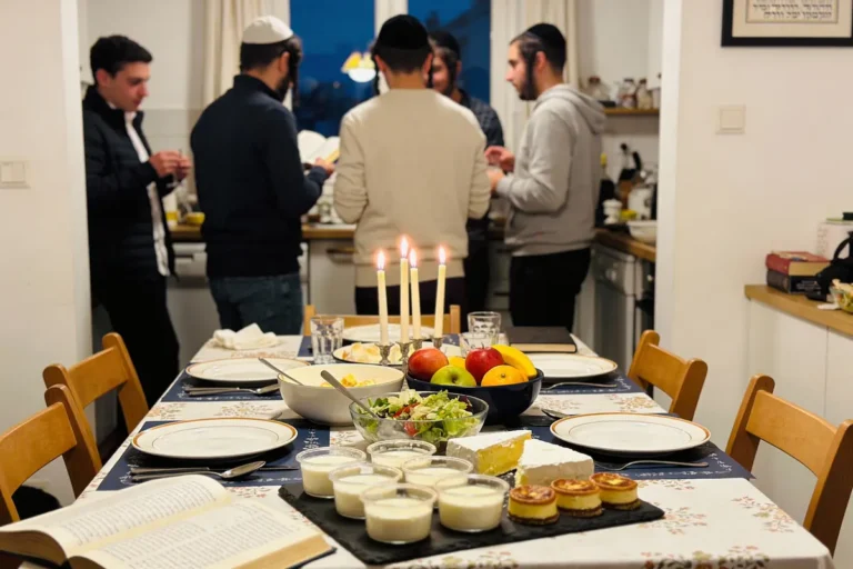 Table dressée avec aliments et chandelles allumées; quatre personnes discutent debout en arrière-plan.