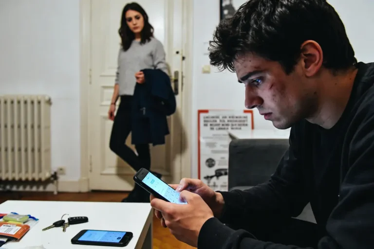Jeune homme concentré sur son téléphone, femme en arrière-plan, atmosphère tendue dans un salon moderne.