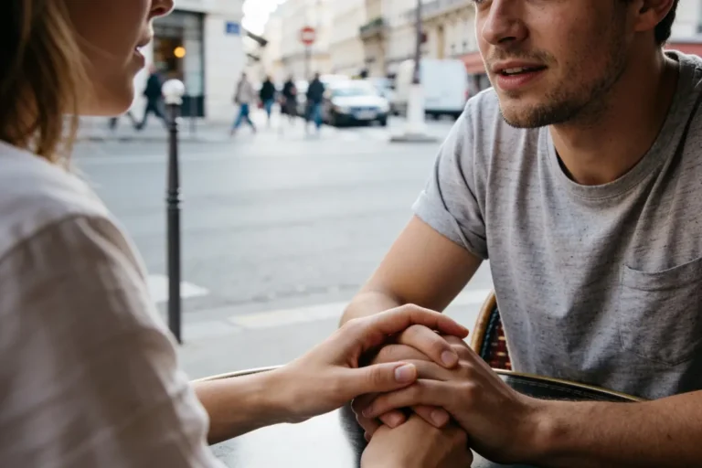 Deux personnes se tiennent la main dans un café en plein air, rue animée en arrière-plan.