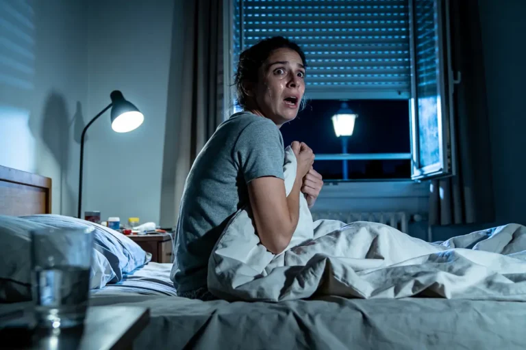 Femme effrayée dans un lit, tenant une couverture, fenêtre ouverte, lampadaire allumé, ambiance nocturne.