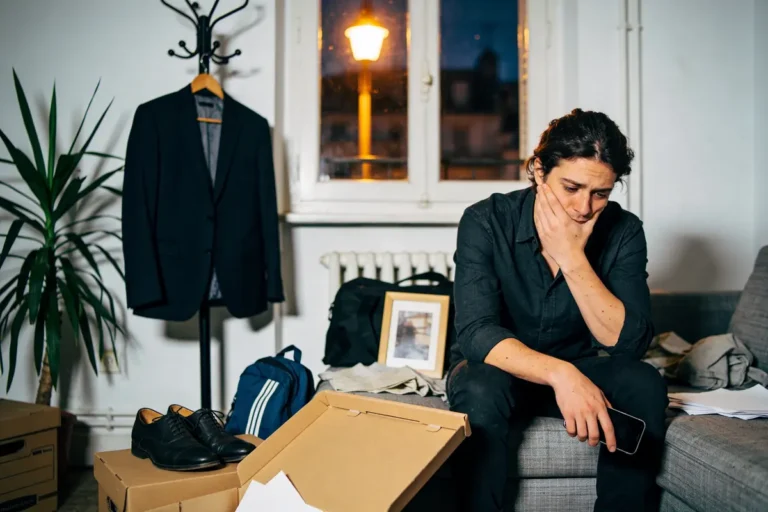 Homme pensif assis sur un canapé entouré de cartons, vêtements et cadre photo, lumière de nuit.