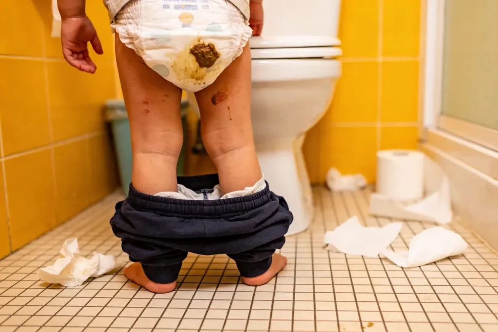 Enfant en couche sale dans une salle de bain, vêtements baissés, papier toilette éparpillé au sol.