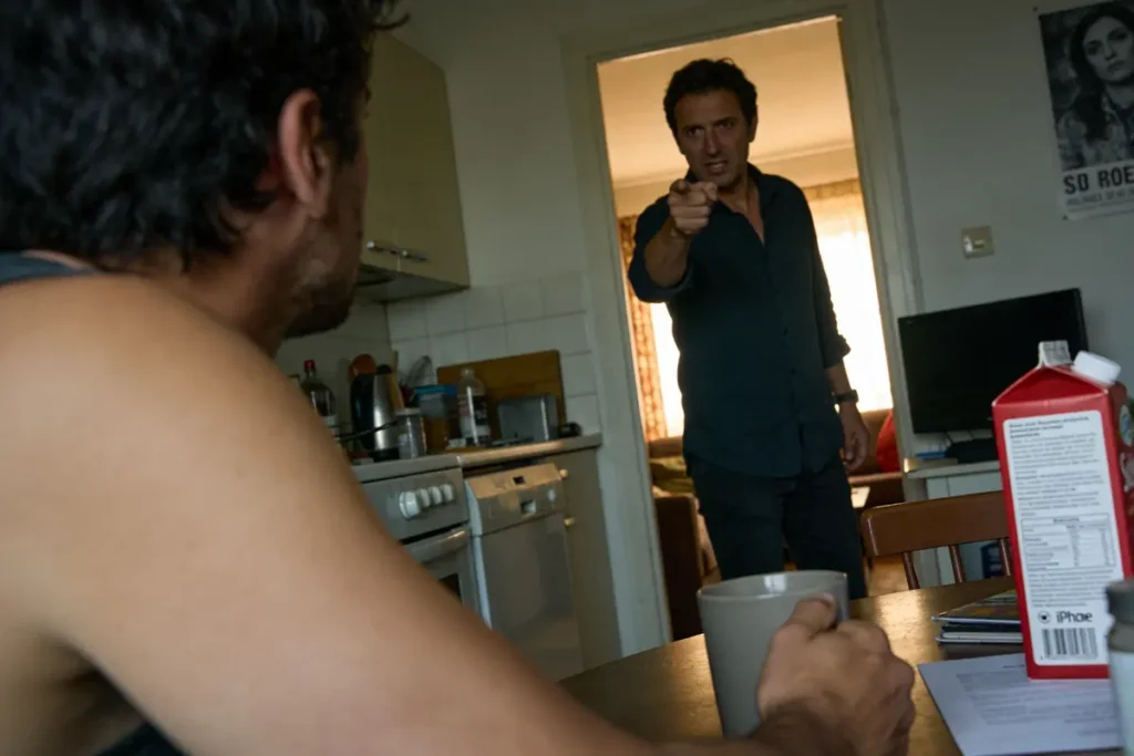 Un homme dans une cuisine, bras tendu, pointant vers une autre personne assise, boîte de lait sur table.