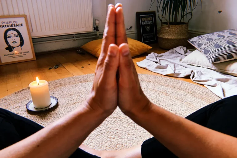 Mains en prière, bougie allumée, tapis rond, ambiance méditative, coussins et affiches en arrière-plan.